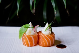 Two pieces of beautifully arranged sushi with a swirl of white creamy topping. The sushi is garnished with green leaves and radish slices, and there is a small pool of dark soy sauce on the white plate. The background has a natural, leafy backdrop that adds to the presentation.