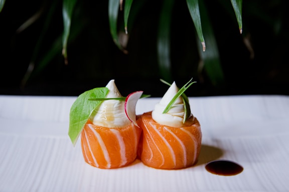 Two pieces of beautifully arranged sushi with a swirl of white creamy topping. The sushi is garnished with green leaves and radish slices, and there is a small pool of dark soy sauce on the white plate. The background has a natural, leafy backdrop that adds to the presentation.