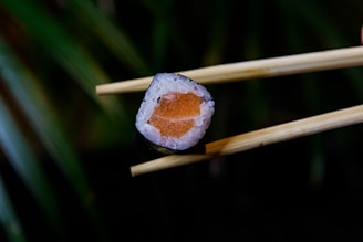 A piece of sushi held by wooden chopsticks, featuring a central piece of salmon surrounded by rice and wrapped in seaweed, set against a blurred background.