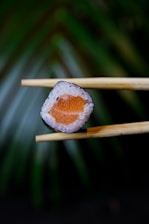 A piece of sushi is delicately held between a pair of wooden chopsticks. The sushi roll features a center filled with bright orange salmon surrounded by white rice. The background consists of a blurred, green leafy texture, creating a smooth bokeh effect.