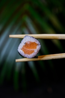 A piece of sushi is delicately held between a pair of wooden chopsticks. The sushi roll features a center filled with bright orange salmon surrounded by white rice. The background consists of a blurred, green leafy texture, creating a smooth bokeh effect.