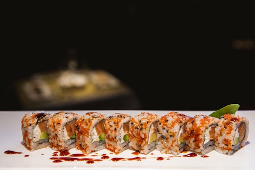 A row of sushi rolls arranged neatly on a white plate, garnished with black sesame seeds and orange tobiko. There is a glossy drizzle of soy sauce on top. A small green leaf is placed at the end of the roll, adding a touch of color.