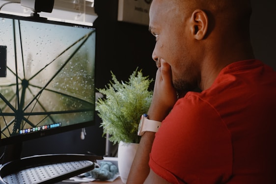 A person in a red shirt is intently looking at a desktop monitor displaying a close-up image of a wet umbrella with raindrops on it. A white smartwatch is on their wrist, and a small green plant is placed on the desk next to the monitor.