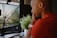 A person in a red shirt is intently looking at a desktop monitor displaying a close-up image of a wet umbrella with raindrops on it. A white smartwatch is on their wrist, and a small green plant is placed on the desk next to the monitor.