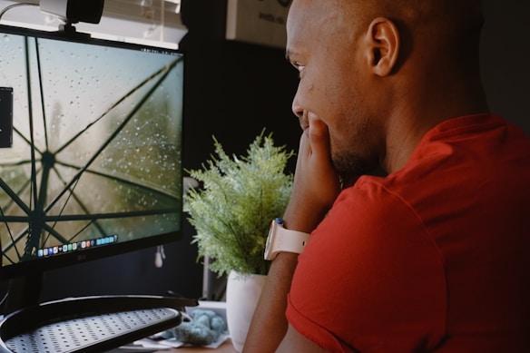 A person in a red shirt is intently looking at a desktop monitor displaying a close-up image of a wet umbrella with raindrops on it. A white smartwatch is on their wrist, and a small green plant is placed on the desk next to the monitor.
