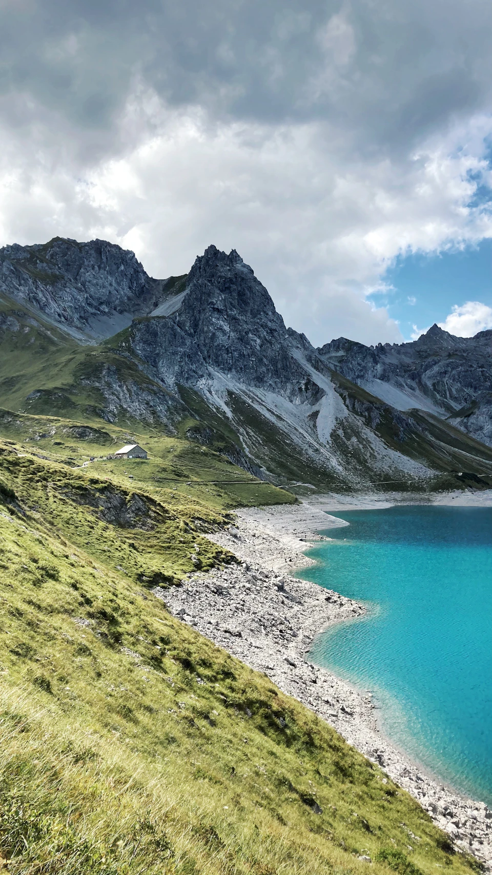 The image depicts a serene mountain landscape with a large, light blue lake nestled at its base. The mountains are covered in lush green vegetation and exhibit varying shades of gray and white, creating a sense of depth and scale. In the foreground, there is a small building partially obscured by the grassy hillside. The sky above is filled with clouds, casting a soft light over the scene and enhancing the overall tranquility of the landscape.
