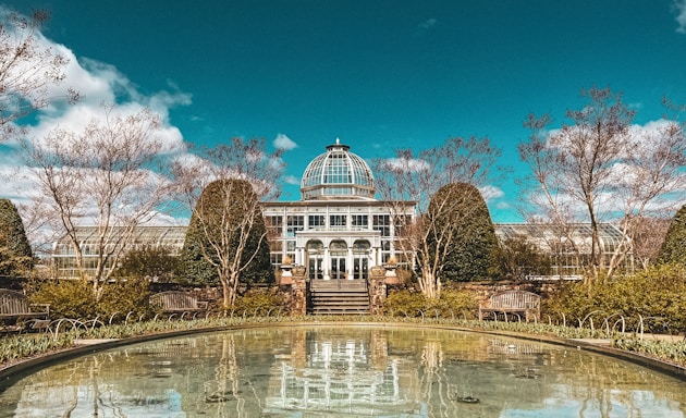 A grand conservatory with a large glass dome is surrounded by well-manicured shrubs and trees. In the foreground, a serene pond reflects the building and the clear blue sky above. Wooden benches are placed symmetrically alongside the water, providing seating.