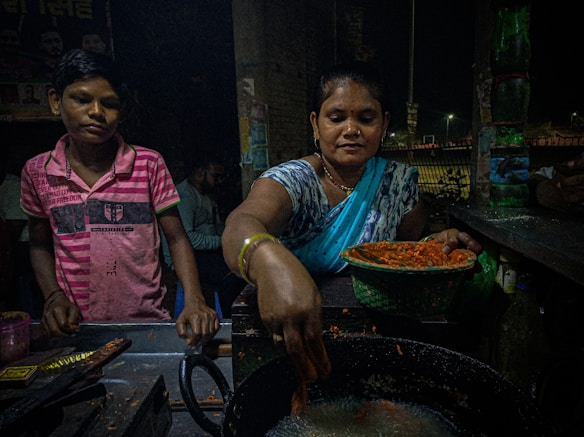 A woman in a blue sari is preparing food at a street stall, placing items into a large pan of oil. A young boy in a pink striped shirt is standing beside her, watching the process. The scene is dimly lit, suggesting it is nighttime, and there are various kitchen items and utensils scattered around.