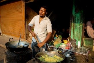 A man is cooking at a street food stall, using a large pan to prepare a dish. Various cooking utensils and ingredients, including tea kettles and plastic bags, are visible on the stall. The setting appears to be an outdoor market with metal shutters and a rustic background.