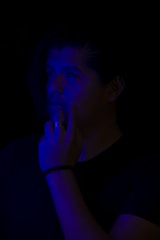 A person is partially illuminated with a blue light, standing against a dark background. Their facial expression suggests deep thought or contemplation, and their hand is raised to their face.