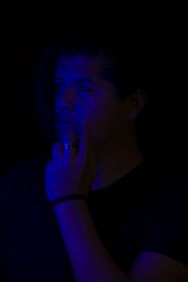 A person is partially illuminated with a blue light, standing against a dark background. Their facial expression suggests deep thought or contemplation, and their hand is raised to their face.