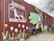 A large mural painted on a brick wall features a white outlined dog above the words 'Brewing Company' and 'Junkyard'. Colorful graphics to the right depict a stylized sun, clouds, and whimsical elements like a train engine and beer glass. Large flowers and stacked wood are present at the bottom of the wall.