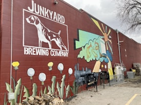A large mural painted on a brick wall features a white outlined dog above the words 'Brewing Company' and 'Junkyard'. Colorful graphics to the right depict a stylized sun, clouds, and whimsical elements like a train engine and beer glass. Large flowers and stacked wood are present at the bottom of the wall.