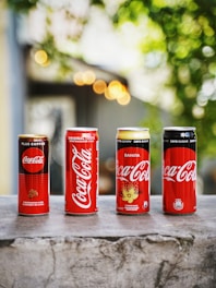 Four different Coca-Cola cans are lined up on a stone surface against a background with bokeh lights and greenery. The cans vary in design and flavor, featuring Plus Coffee, Original Taste, Vanilla, and Zero Sugar options.
