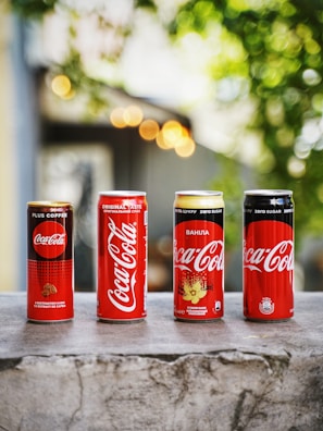 Four different Coca-Cola cans are lined up on a stone surface against a background with bokeh lights and greenery. The cans vary in design and flavor, featuring Plus Coffee, Original Taste, Vanilla, and Zero Sugar options.