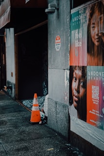 A dimly lit urban street scene, featuring a concrete wall with posters advertising beauty products. A no trespassing sign is visible above the posters. A traffic cone and a makeshift bedding setup are placed on the sidewalk, suggesting urban life and possibly homelessness.