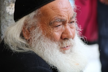 An elderly man with a long white beard and a contemplative expression. He is wearing a black hat and dark clothing, with deep facial wrinkles that suggest wisdom and experience. The background is softly blurred with hints of red and grey.