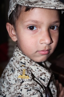 A young child wearing a military-style camouflage uniform and cap, with a focused expression on their face. The uniform features a detailed pattern with an insignia on the shoulder.