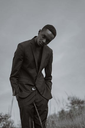 A person in a dark suit standing in an outdoor setting with a slightly tilted posture. The background is a muted, cloudy sky with some grass and foliage visible in the lower portion.