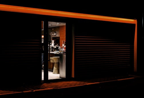 A dimly lit establishment with a warm, inviting interior is visible through an open door. The exterior is dark and sleek, accented by an orange strip of light at the top of the entrance. Inside, modern furniture, including a wicker chair and a counter, are observable, along with some hanging lights and a menu display.