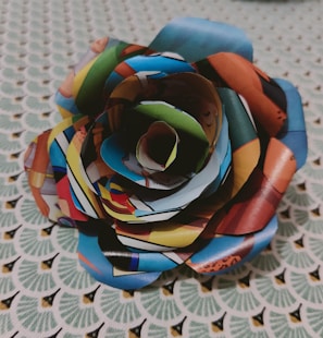 A flower crafted from folded and cut colorful paper sits on a textured background. The paper appears to be sourced from a magazine or comic, showcasing various vivid images and text in a collage style. The background is a patterned fabric with a repeating geometric design in green and white.
