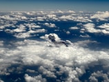 Clouds spread across the sky, with a plume of smoke or ash emerging from a darker area among the white formations. The view is from above, showcasing an expansive and dramatic skyscape.