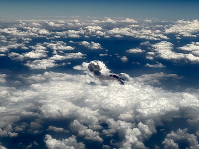 Clouds spread across the sky, with a plume of smoke or ash emerging from a darker area among the white formations. The view is from above, showcasing an expansive and dramatic skyscape.