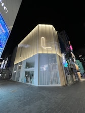 A modern, illuminated building on a nighttime street corner. The building features sleek architecture with translucent panels and a prominent 'L' logo. The surrounding area appears quiet, with a few other buildings visible, including signs with illuminated text.