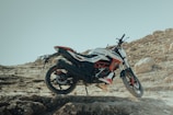 A dirt bike is parked on a rugged, rocky terrain with a clear blue sky in the background. The bike features a sporty design with a color scheme that includes black, white, and red. Large rocks and uneven ground characterize the setting, emphasizing a sense of adventure and off-road exploration.