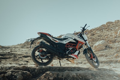 A dirt bike is parked on a rugged, rocky terrain with a clear blue sky in the background. The bike features a sporty design with a color scheme that includes black, white, and red. Large rocks and uneven ground characterize the setting, emphasizing a sense of adventure and off-road exploration.