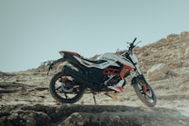 A dirt bike is parked on a rugged, rocky terrain with a clear blue sky in the background. The bike features a sporty design with a color scheme that includes black, white, and red. Large rocks and uneven ground characterize the setting, emphasizing a sense of adventure and off-road exploration.