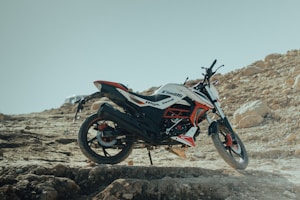 A dirt bike is parked on a rugged, rocky terrain with a clear blue sky in the background. The bike features a sporty design with a color scheme that includes black, white, and red. Large rocks and uneven ground characterize the setting, emphasizing a sense of adventure and off-road exploration.