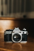 A vintage Canon AE-1 film camera is positioned on a wooden surface. The camera body is prominently displayed with its lens mount exposed. The scene is warmly lit, highlighting the metallic and textured black elements of the camera.