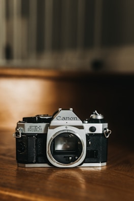 A vintage Canon AE-1 film camera is positioned on a wooden surface. The camera body is prominently displayed with its lens mount exposed. The scene is warmly lit, highlighting the metallic and textured black elements of the camera.