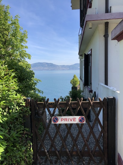 A wooden gate labeled 'PRIVÉ' in front of a gravel path, leading to a scenic view of a large body of water with distant mountains. The gate is surrounded by lush greenery on the left and a building wall on the right. The sky is clear and blue.
