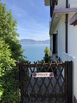 A wooden gate labeled 'PRIV&Eacute;' in front of a gravel path, leading to a scenic view of a large body of water with distant mountains. The gate is surrounded by lush greenery on the left and a building wall on the right. The sky is clear and blue.