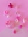 An assortment of small, colorful craft beads arranged on a pink surface. The collection includes shapes such as hearts, stars, and a strawberry with polka dots. The beads vary in shades of pink, purple, and white, creating a playful and cheerful composition.