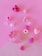 An assortment of small, colorful craft beads arranged on a pink surface. The collection includes shapes such as hearts, stars, and a strawberry with polka dots. The beads vary in shades of pink, purple, and white, creating a playful and cheerful composition.