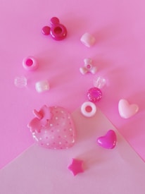 An assortment of small, colorful craft beads arranged on a pink surface. The collection includes shapes such as hearts, stars, and a strawberry with polka dots. The beads vary in shades of pink, purple, and white, creating a playful and cheerful composition.