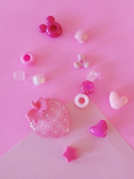 An assortment of small, colorful craft beads arranged on a pink surface. The collection includes shapes such as hearts, stars, and a strawberry with polka dots. The beads vary in shades of pink, purple, and white, creating a playful and cheerful composition.