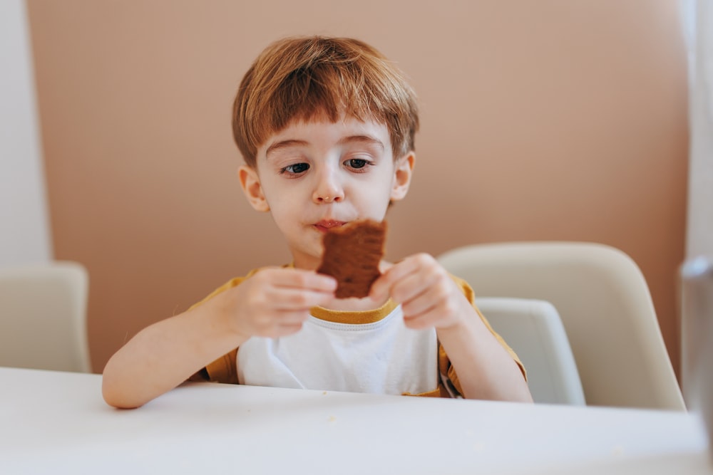 a young boy sitting at a table eating a piece of food