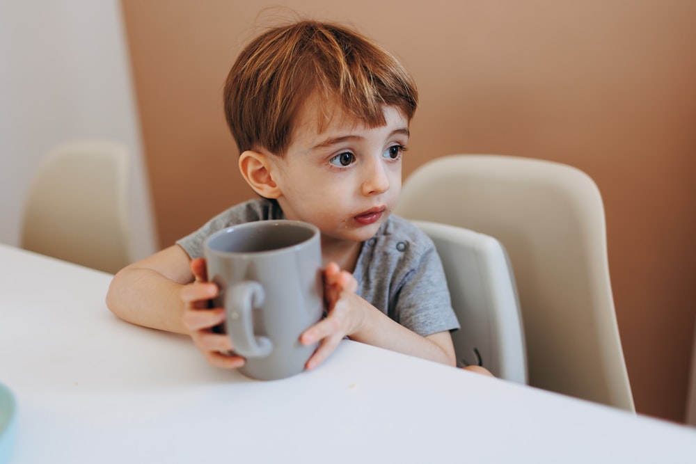 a little boy sitting at a table holding a cup