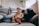 A woman and a child are comfortably sitting on a sofa at home. The child is holding a remote control, and both are smiling, conveying a sense of relaxation and happiness. There are plants in the background, adding a touch of greenery to the cozy living space.