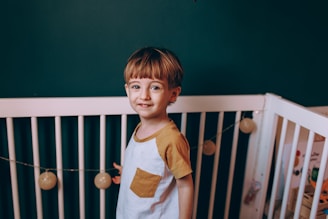 A young child stands inside a white crib against a dark green wall. The child is wearing a white shirt with mustard yellow sleeves and pocket. There are decorative string lights with round bulbs hanging across the crib.