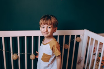 A young child stands inside a white crib against a dark green wall. The child is wearing a white shirt with mustard yellow sleeves and pocket. There are decorative string lights with round bulbs hanging across the crib.