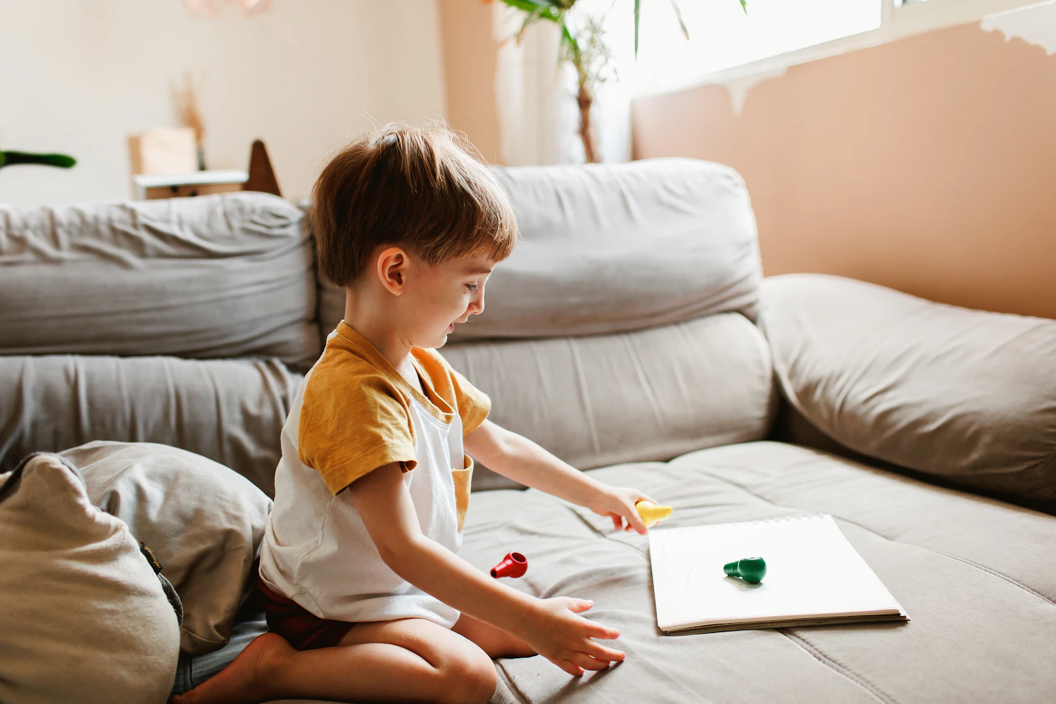 A little boy playing calmly with crayons and a sketchbook on the couch at home