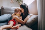A woman is sitting on a couch, lovingly hugging and kissing a young child who is sitting on her lap. The setting is cozy, with soft cushions and warm light filtering through a window, creating a serene and intimate atmosphere.