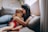 A woman is sitting on a couch, lovingly hugging and kissing a young child who is sitting on her lap. The setting is cozy, with soft cushions and warm light filtering through a window, creating a serene and intimate atmosphere.