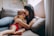 A woman is sitting on a couch, lovingly hugging and kissing a young child who is sitting on her lap. The setting is cozy, with soft cushions and warm light filtering through a window, creating a serene and intimate atmosphere.