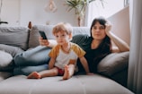 A woman and a young boy are sitting together on a comfortable gray couch in a living room. The woman is leaning back, appearing relaxed, with her hand resting on her head. The boy is sitting in front of her, looking attentively forward as the woman holds a TV remote. A large potted plant and a window with sheer curtains are visible in the background, adding a cozy ambiance to the scene.